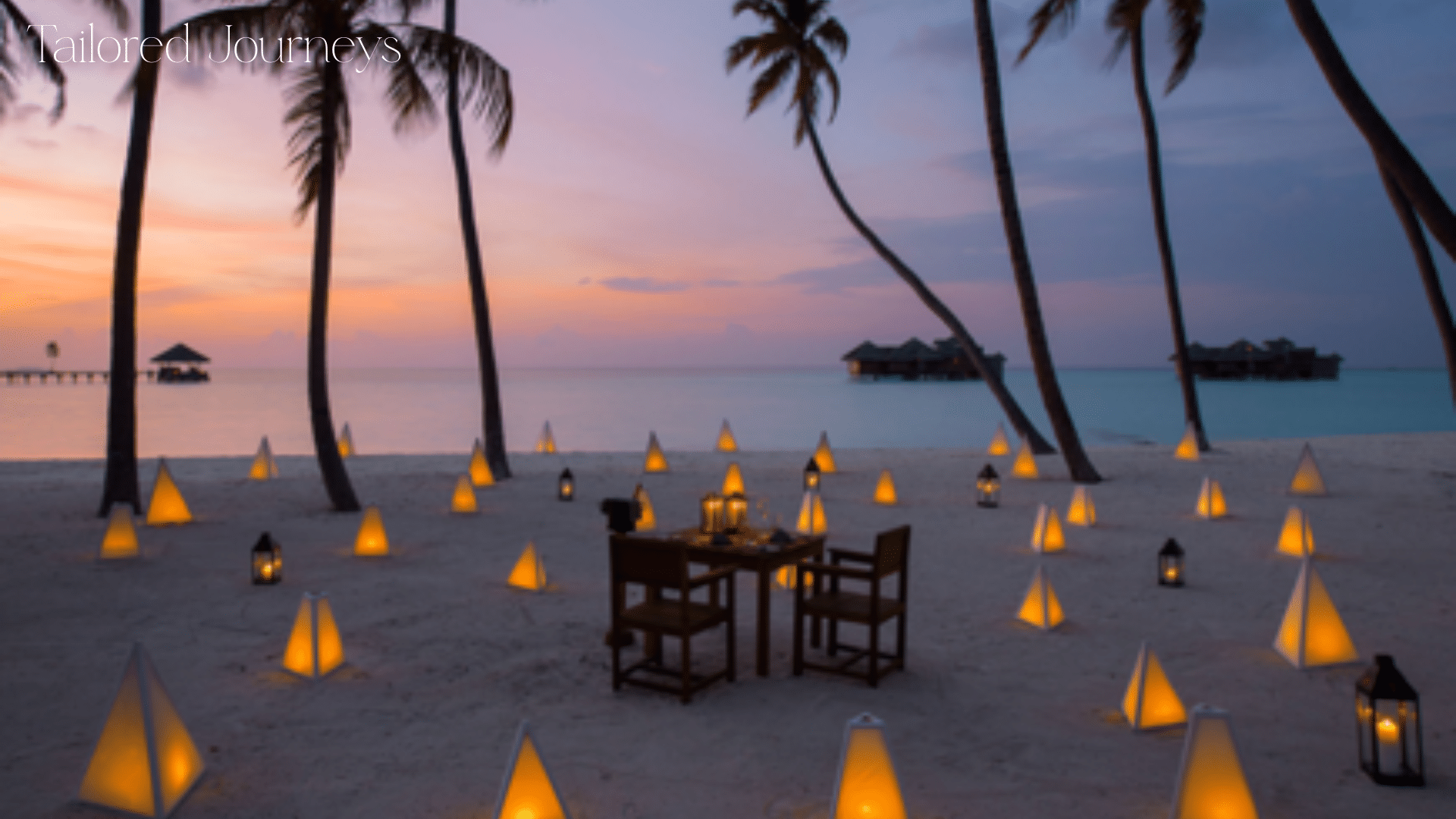 A romantic beach scene, a table with two chairs on a beach surrounded by lanterns with the sun setting