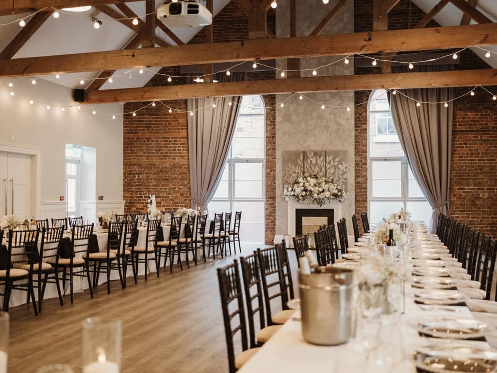 Elegant wedding reception setup in a luxury, affordable Kent wedding venue, featuring long banquet tables, exposed brick walls, wooden beams, and soft string lighting.