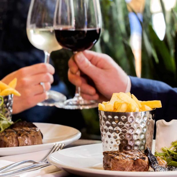 Two people clinking wine glasses over a delicious steak dish, enjoying Black Friday at The Brasserie